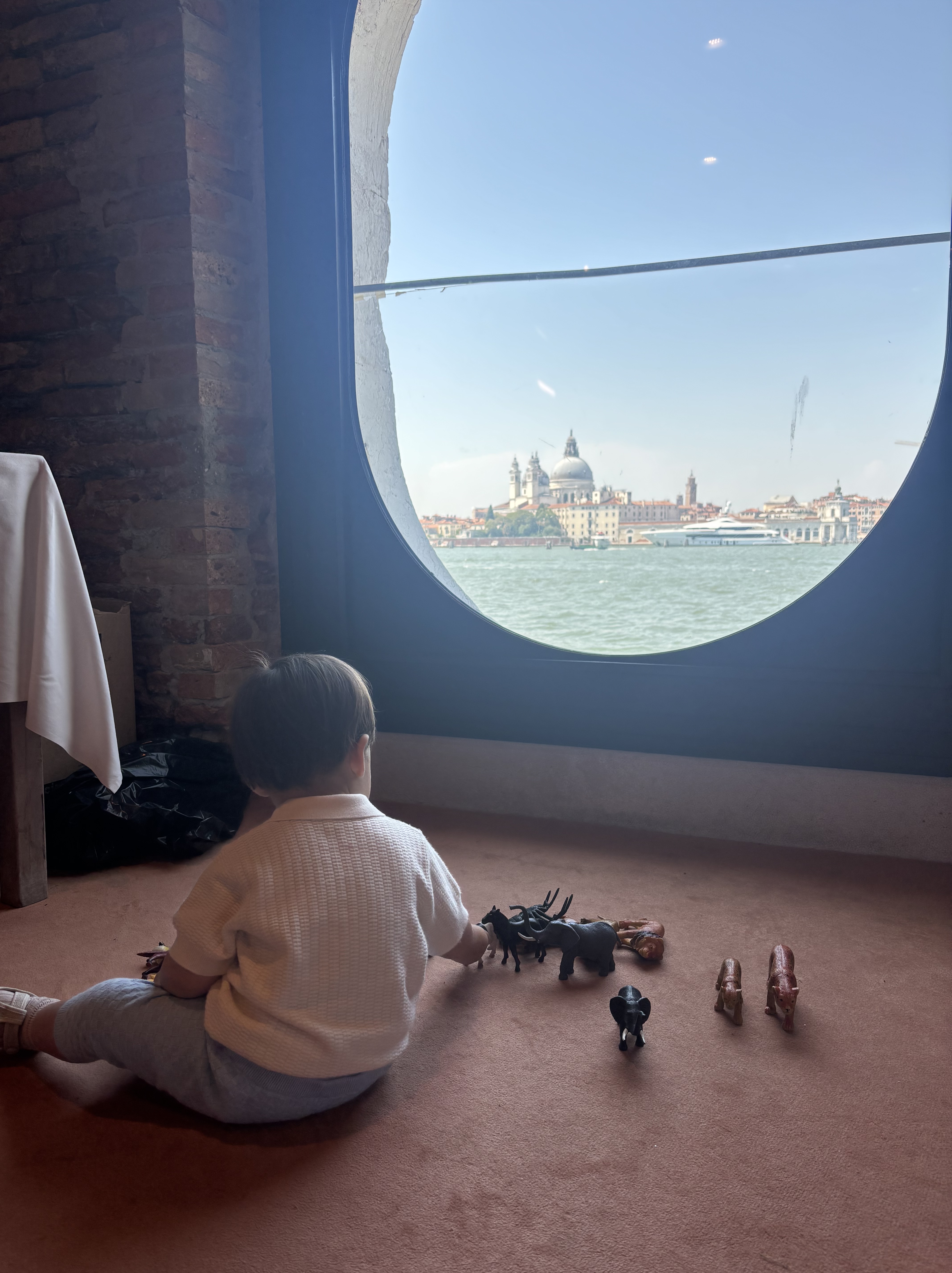 A meticulously arranged luxury babysitting setup in a spacious, modern hotel suite in Venice, photographed in realistic detail. A plush, cream-colored play rug lies on polished oak parquet, hosting a small collection of high-quality wooden toys, linen picture books, and a neatly folded cashmere baby blanket in soft dove grey. In the background, a large window reveals an elegant, softly blurred view of Venetian rooftops and a hint of a canal. Warm late-afternoon sunlight pours in, casting gentle, elongated shadows and subtle highlights on metallic toy details. Shot at eye level with a shallow depth of field, the composition feels calm, refined, and welcoming, embodying a sophisticated, five-star aesthetic suitable for discerning families.